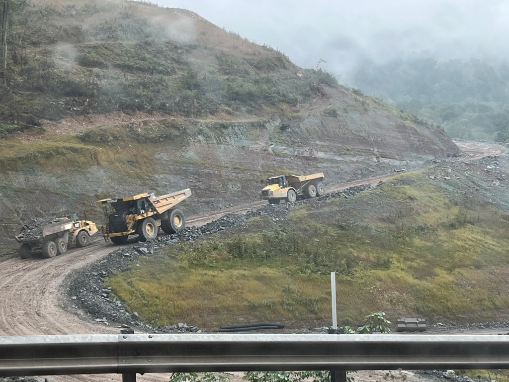 Two large trucks transporting gravel on a muddy construction site surrounded by hilly terrain and foggy weather.