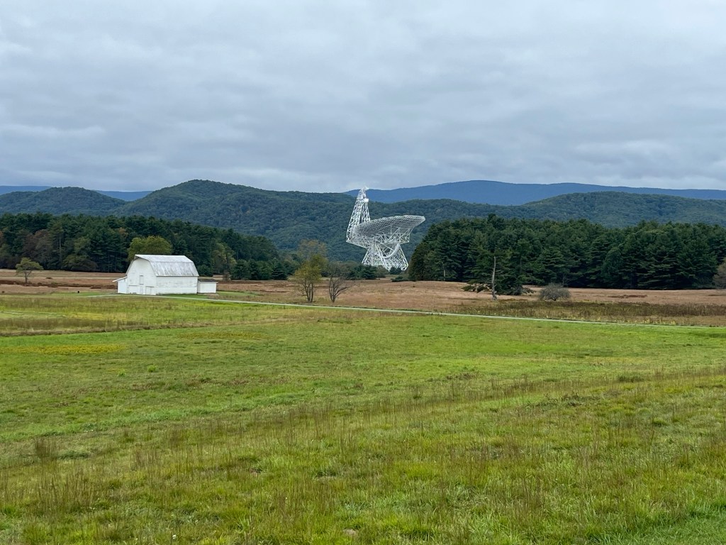 A white barn in a green field with a large radio telescope structure in the background, surrounded by rolling hills and cloudy skies.