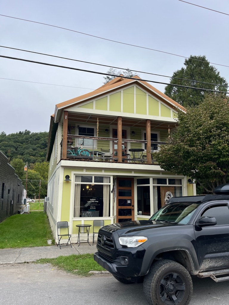 A two-story yellow building with a porch and outdoor seating, featuring large windows and a black vehicle parked in front.