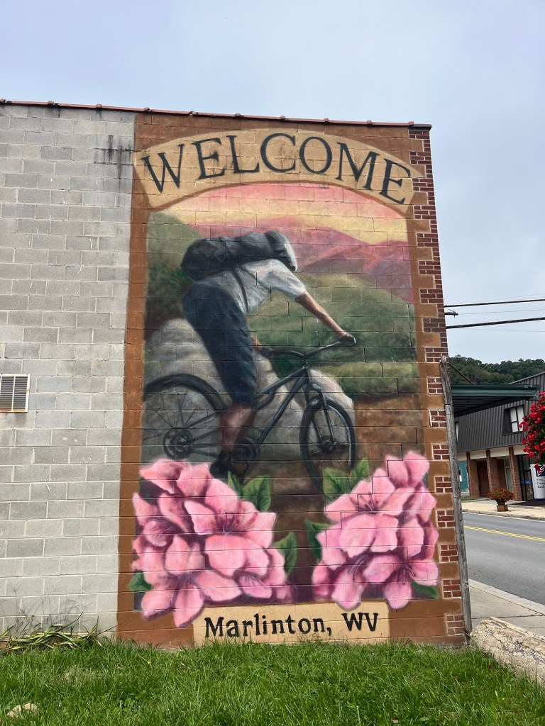 A mural featuring the word 'WELCOME' at the top, depicting a mountain biker riding through a scenic landscape with pink flowers at the bottom. The mural is located in Marlinton, WV.