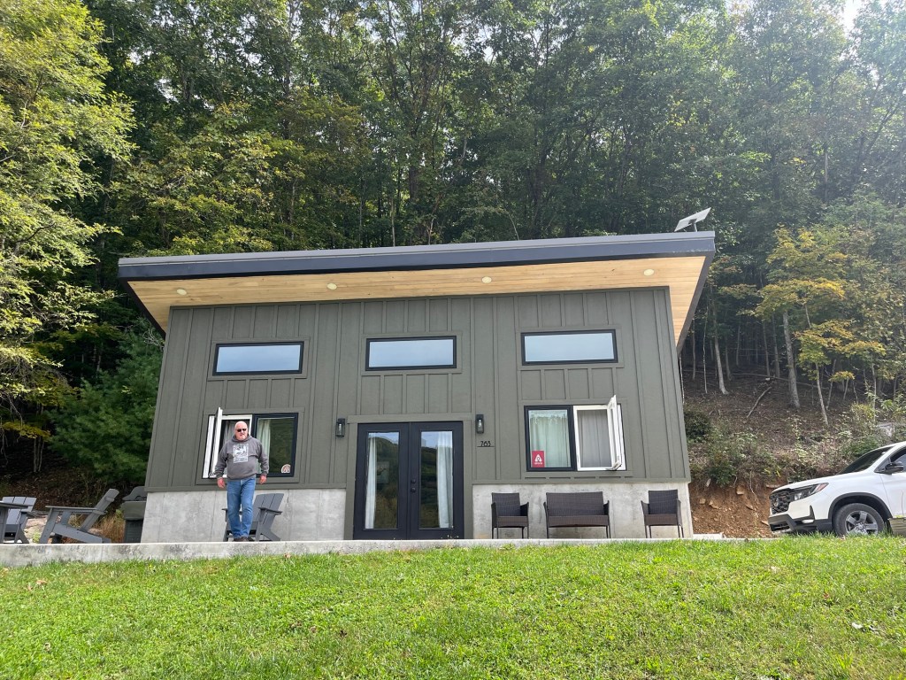 A modern green cabin with large windows, a flat roof, and a man standing in front. Surrounding trees provide a natural backdrop.