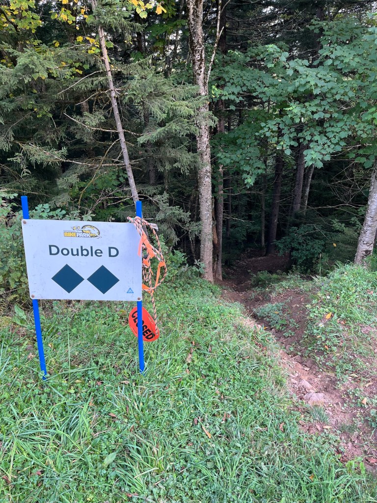 Sign marking the trail 'Double D' at a bike park, with a closed sign and forested area in the background.