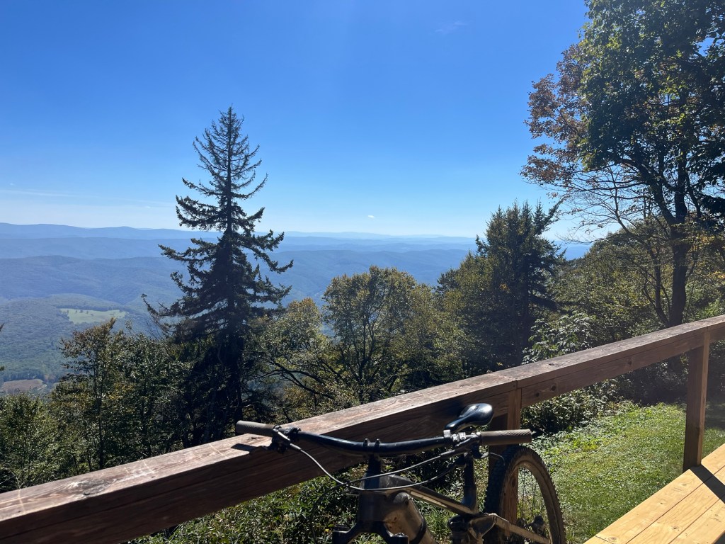 A scenic view from a mountain overlook featuring a wooden railing, with a mountain bike leaning against it. In the background, rolling mountains and a clear blue sky are visible, surrounded by trees and greenery.