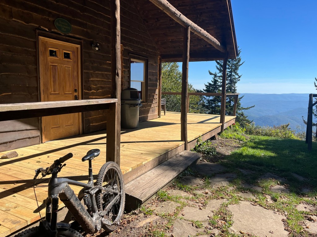 A rustic log cabin with a wooden porch overlooks a scenic view of mountains under a clear blue sky. A mountain bike is leaning against the porch railing.