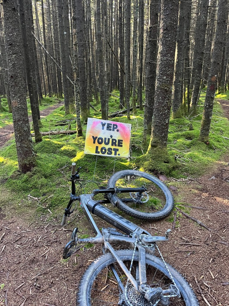 A black bicycle resting on the ground in a forest, next to a colorful sign that says 'Yep, You're Lost.' The background features tall trees and moss-covered ground.